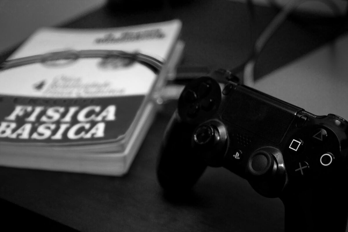 Fotografia em preto em branco de um livro no qual se lê "física básica 4", sobre uma mesa ao lado de um controle de Playstation 4. Há cabos de conexão que saem do livro.